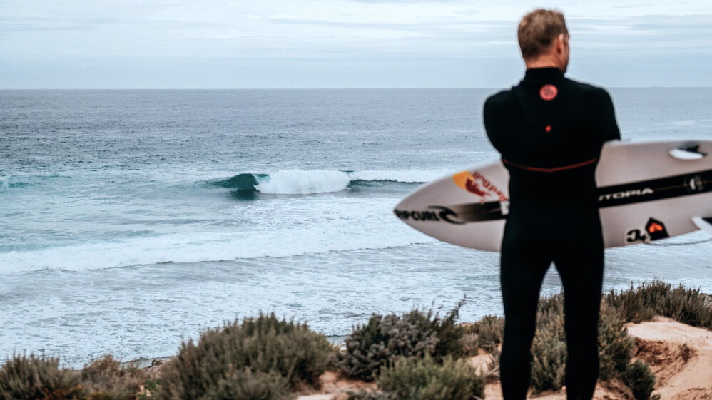 Mann steht mit seinem Surfbrett auf einer Klippe.